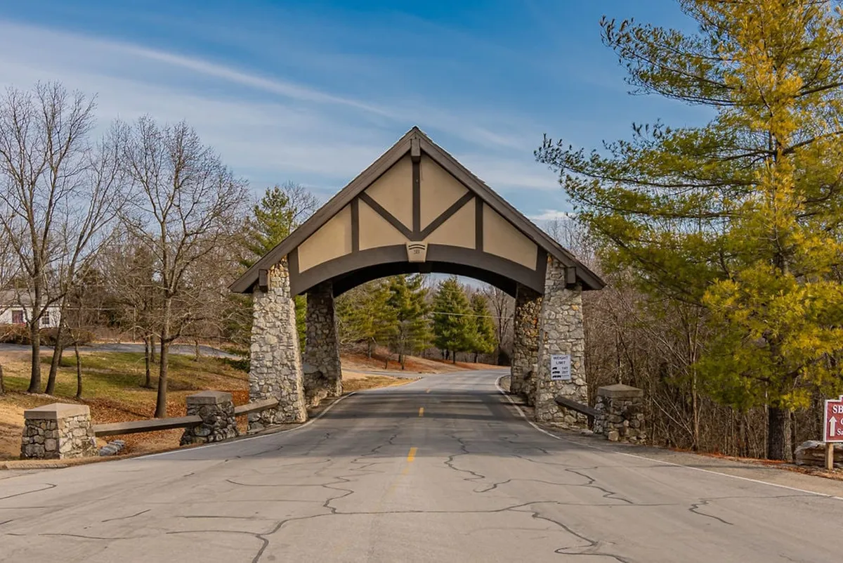 This exterior shot features a prominent stone archway marking the gated entrance to the community, surrounded by mature trees and a quiet, tree-lined road. Signage indicates the private nature of the property, giving a sense of exclusivity and security.