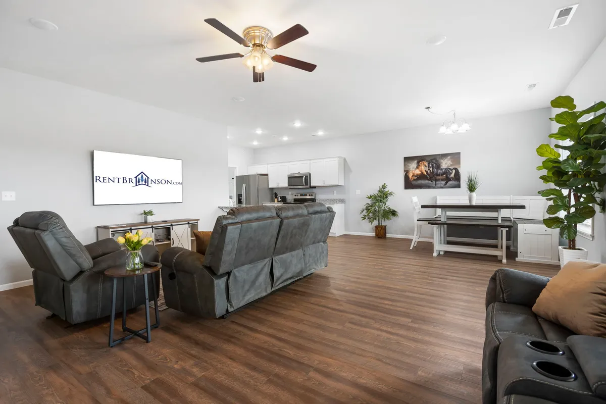 A wide view of the living/dining space displays comfortable seating, a large TV, a dining table set for meals, potted plants, and tasteful artwork. The space is arranged to facilitate both relaxation and shared experiences, blending entertainment and dining seamlessly.