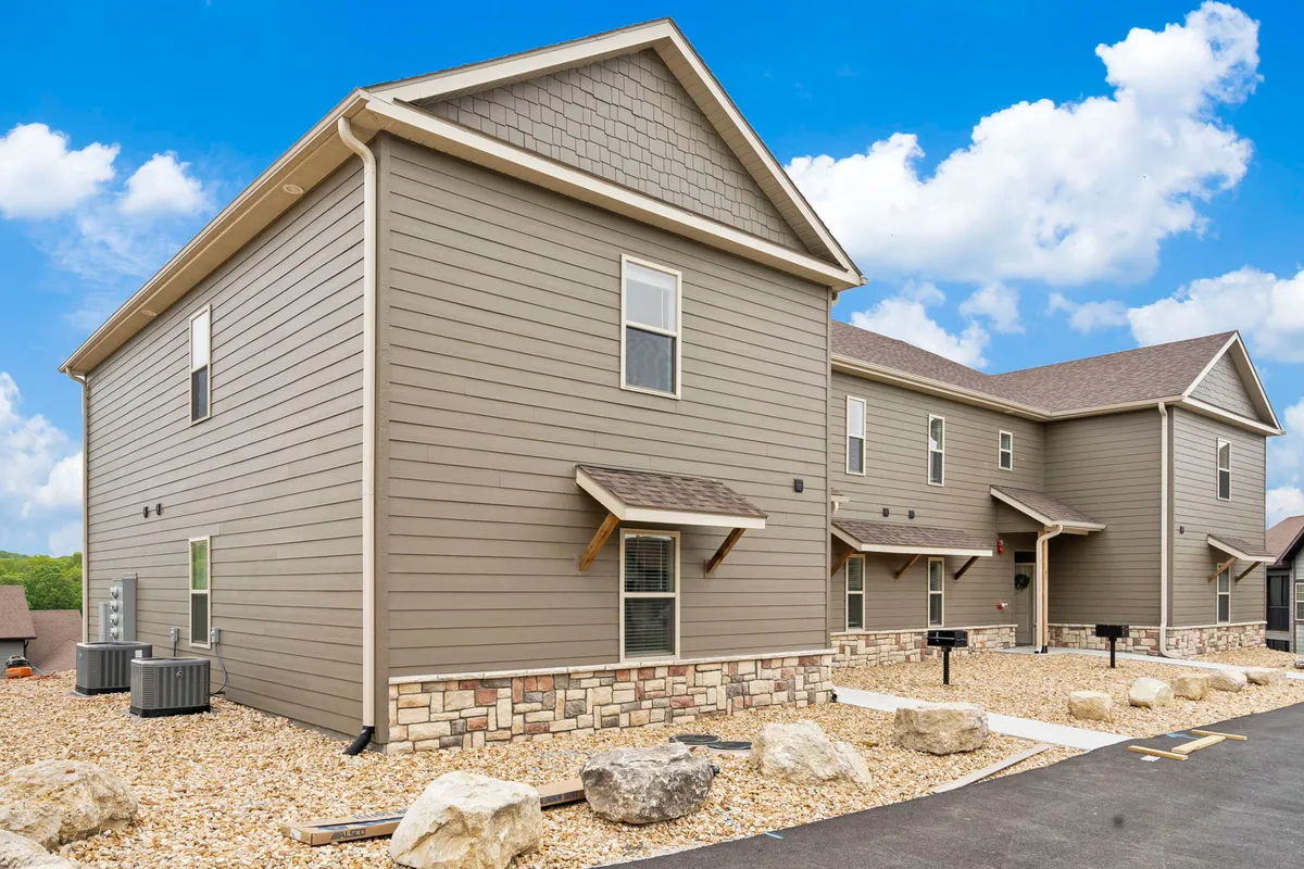 This exterior view showcases modern architecture with stone accents, a neatly paved driveway, and beautifully landscaped rocks. The welcoming tone, combined with the picturesque surroundings, conveys a sense of security, comfort, and prestige within a gated community environment.