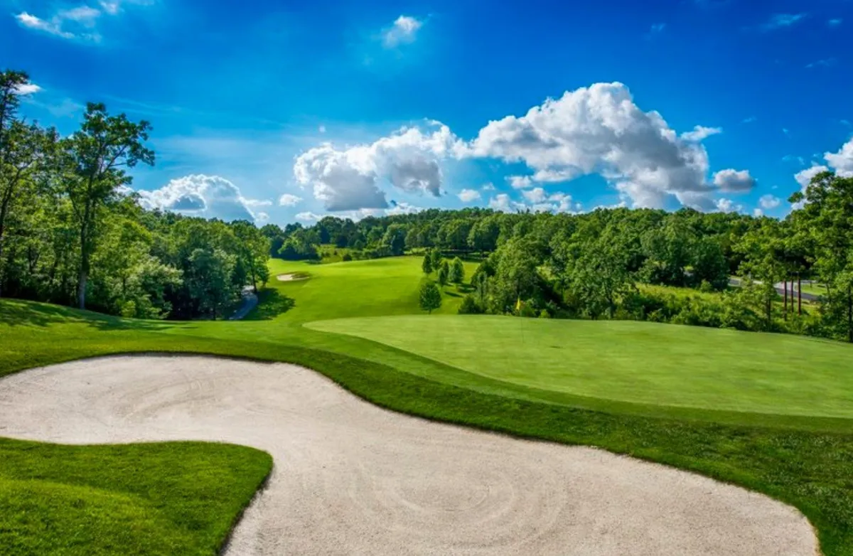 This image captures a lush golf course with a sand trap, vibrant green fairway, distant trees, and a blue sky scattered with clouds. The landscape is open and inviting, embodying the setting for a memorable round of golf.
