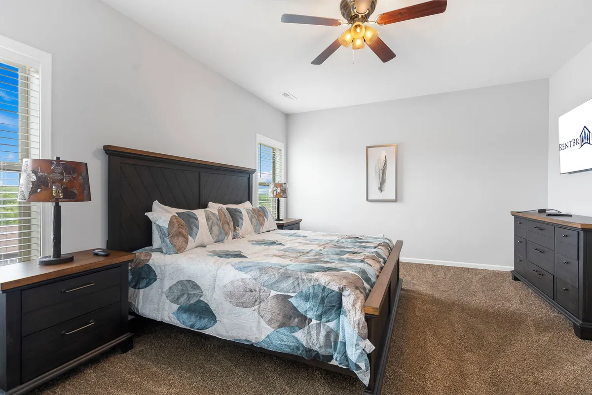 A serene bedroom with a large, inviting bed dressed in patterned bedding, a nightstand with a lamp for nighttime reading, a wall-mounted TV for entertainment, and framed art adding a touch of modern decor. Soft natural light filters through nearby windows, enhancing the tranquil atmosphere.
