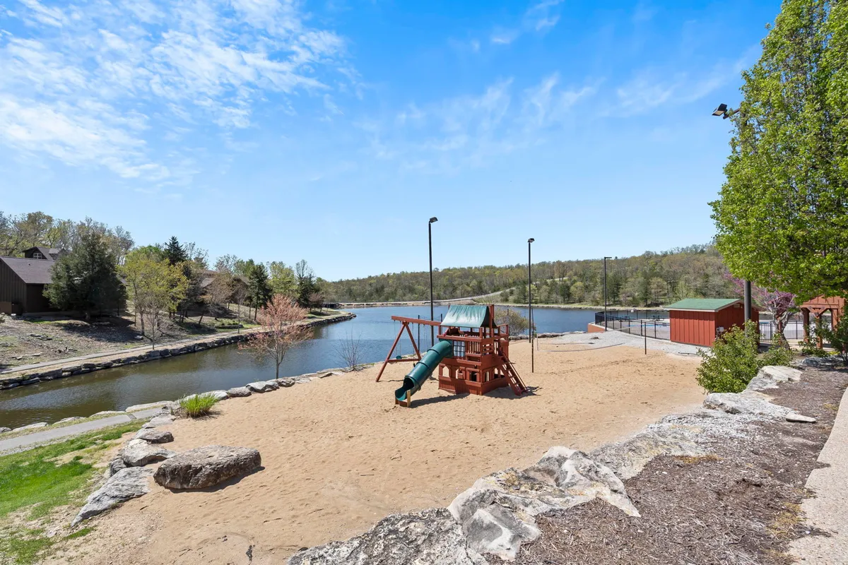 A vibrant playground with slides and swings set beside a scenic lake and walking path, surrounded by trees. The space is ideal for families with children, offering opportunities for play, picnicking, and lakeside relaxation.
