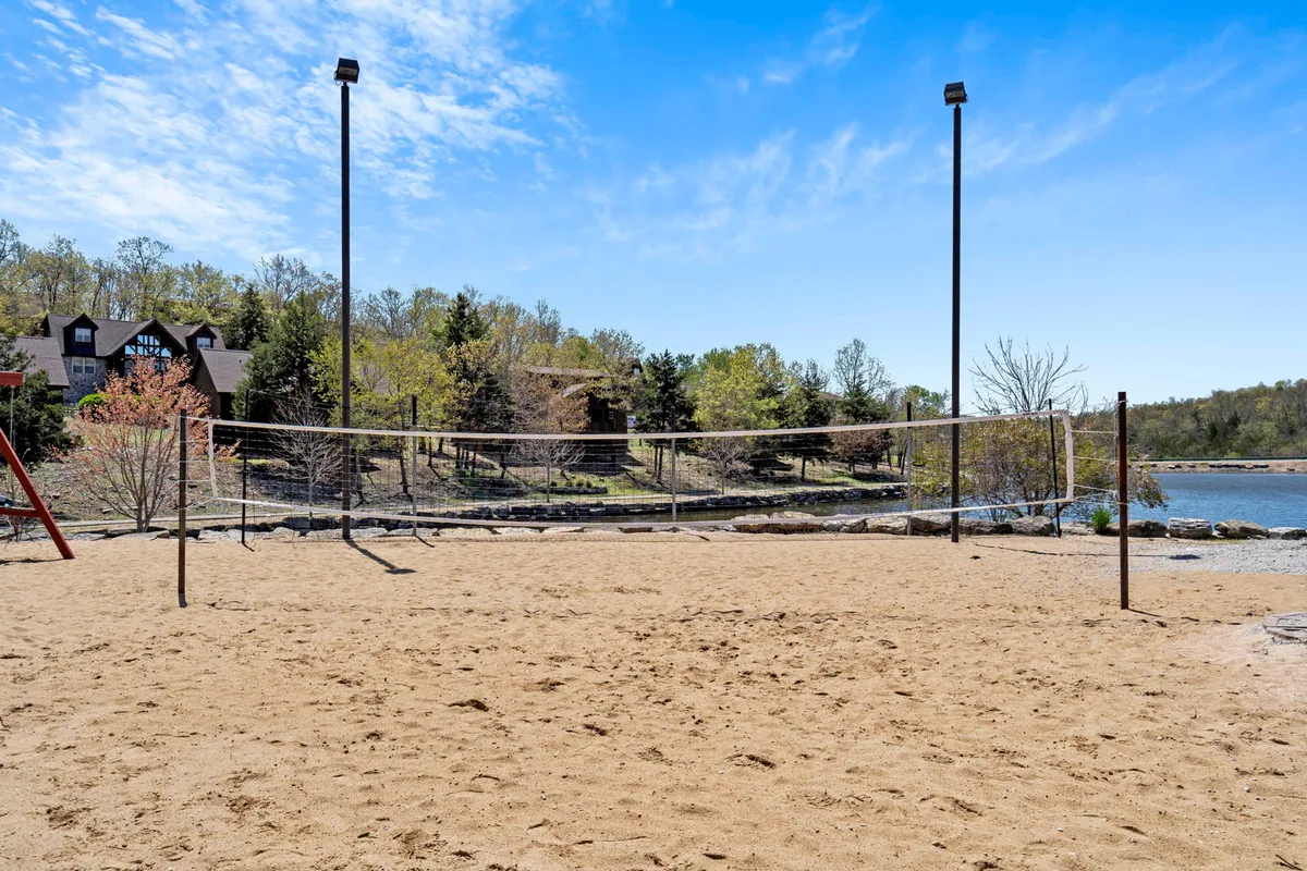 This scene showcases a sand volleyball court set by a picturesque lake, surrounded by trees and a background building. The bright sand, net, and adjacent nature hint at both energetic play and lakeside relaxation, perfect for group gatherings and friendly competition.