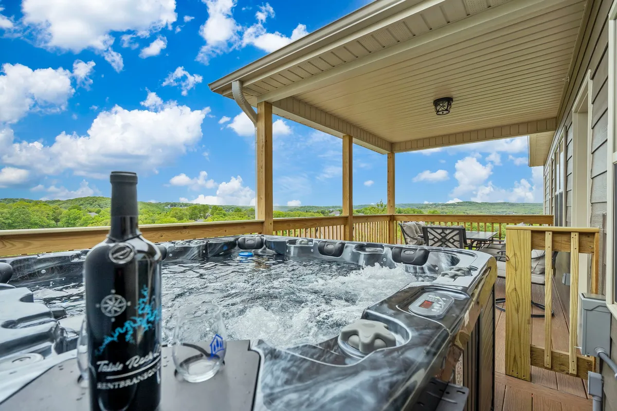 The image shows a private outdoor hot tub on the condo's patio, with a bottle of wine set out for guests. The hot tub overlooks rolling Ozark hills, and the scene is set at sunset, creating a tranquil, inviting atmosphere ideal for relaxation and socializing.