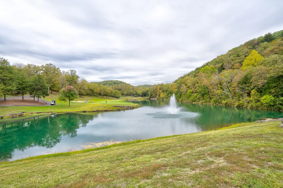 This serene view features a calm lake with a fountain, tree-lined shore, and rolling hills in the distance. The tranquil setting is ideal for fishing, picnicking, or simply relaxing and enjoying nature's beauty.