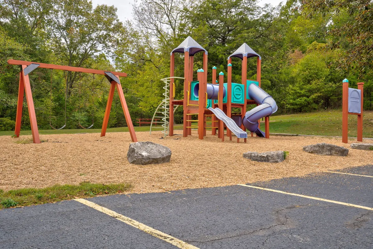 The image showcases a joyful playground scene with play equipment, swings, and rock features, set in a safe and inviting outdoor environment. Children are pictured playing, emphasizing laughter and energetic family interaction.